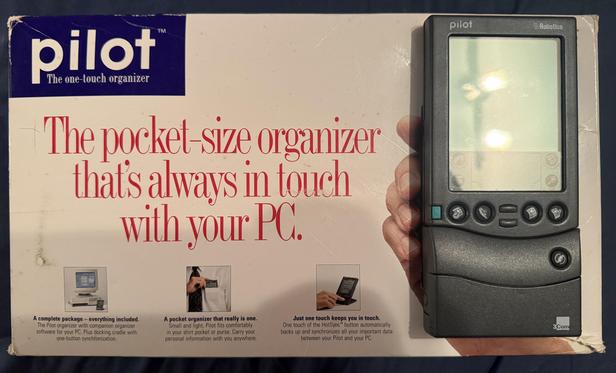 A Pilot 1000 PDA, a handheld touchscreen organizer from the 1990s, with a PalmPilot Modem attached to the bottom. It is sitting on top of the Pilot’s box, on top of where a hand is holding a photo of the device