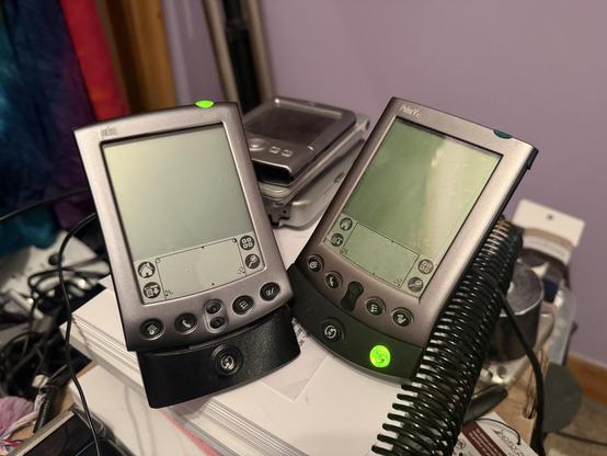 A Palm m500 (left) on its charging dock, beside a Palm Vx (right) on its dock, both thin silver PDA devices. In the background a Palm Tungsten T2 is visible, on top of a Palm LifeDrive and a Sony Clie NX70