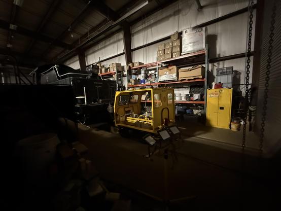The small yellow wooden railway vehicle, in the railway shop with most of the lights off. A coal-carrying tender sits on the same track just ahead of it